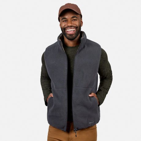 Smiling man wearing a brown cap, dark green long-sleeve shirt, brown pants, and the Free Country Men’s Haven Stitchless Reversible Vest in gray for lightweight warmth, standing with hands in vest pockets against a plain white background.