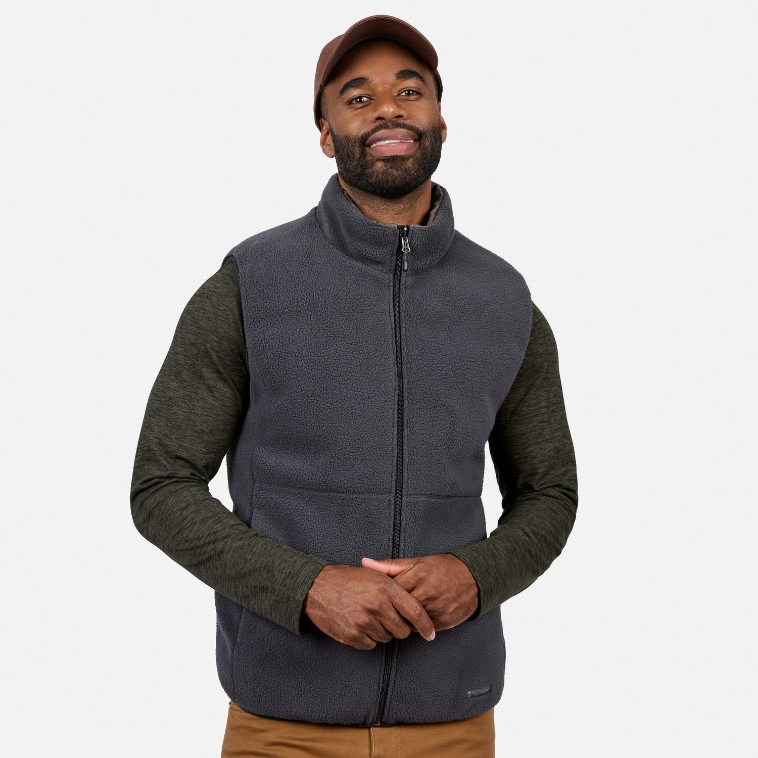 A man wearing a brown cap, dark green long-sleeve shirt, and brown pants stands against a plain white background, smiling with his hands loosely clasped in front while modeling the Free Country Men’s Haven Stitchless Reversible Vest in gray.