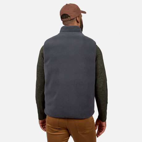 A man is shown from the back against a plain white background wearing the Free Country Men’s Haven Stitchless Reversible Vest in a dark color, paired with an olive long-sleeve shirt, brown pants, and a brown baseball cap.