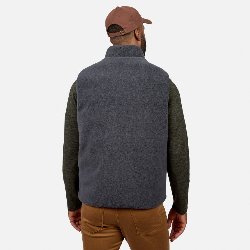 A man is shown from the back against a plain white background wearing the Free Country Men’s Haven Stitchless Reversible Vest in a dark color, paired with an olive long-sleeve shirt, brown pants, and a brown baseball cap.