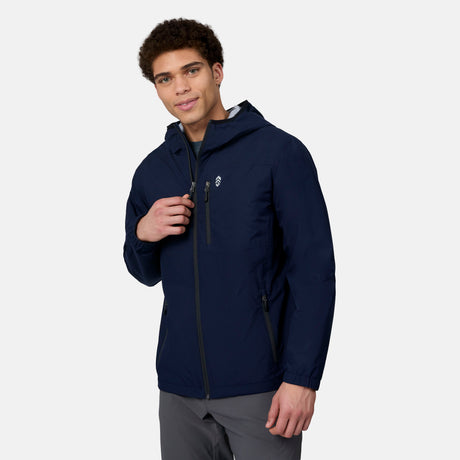 A man wears the Free Country Men's Agile II Windshear Jacket, a dark blue, lightweight, wind-resistant coat with zippered pockets, standing against a plain white background while looking at the camera and smiling slightly.