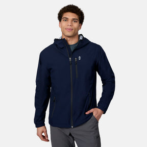 A man stands against a light gray background, smiling slightly with one hand in his pocket, wearing the Free Country Men's Agile II Windshear Jacket in navy blue—a lightweight, water-resistant coat with zipper pockets.