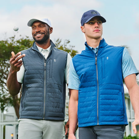 Two men wear Clearance Men's High Stretch Full Zip Vests—one dark gray, one bright blue with recycled polyfill—over polo shirts and baseball caps. The man on the left smiles and tosses a golf ball; the other gazes ahead. Trees and buildings are behind them.