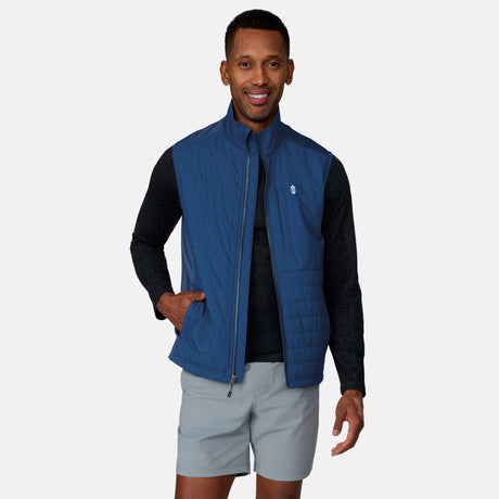 A man smiles while wearing the Clearance Men's High Stretch Full Zip Vest over a black long-sleeve shirt and light gray shorts. One hand is in his vest pocket, against a plain, light-colored background.