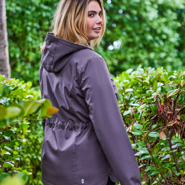 A woman with shoulder-length blonde hair stands outdoors among green bushes and trees, wearing the Free Country Women's Plus Size X2O Anorak Rain Jacket in gray and looking over her shoulder.