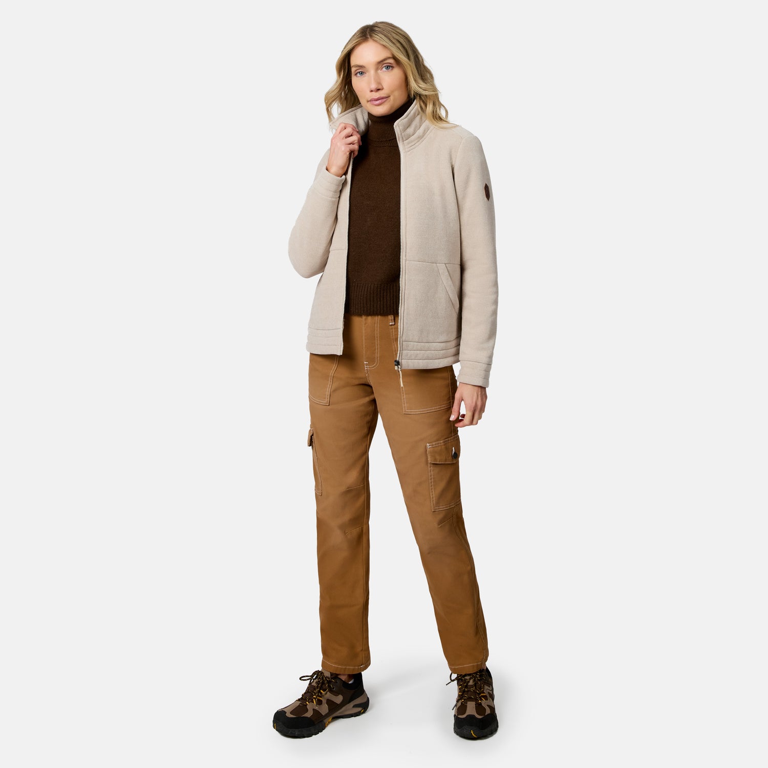 A woman modeling a light beige fleece jacket over a dark brown turtleneck, paired with brown cargo pants and brown hiking boots, against a plain light gray studio background.