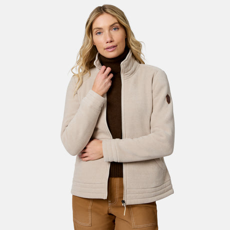 A woman modeling a light beige fleece jacket with a stand-up collar, worn over a dark brown turtleneck and tan pants, against a plain light gray background.