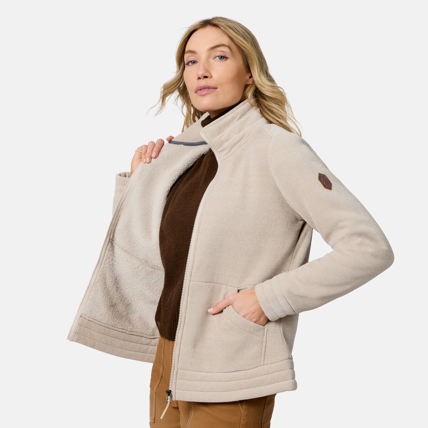 A woman wearing a light beige fleece jacket with a brown mock neck underneath, posed against a solid light gray background.