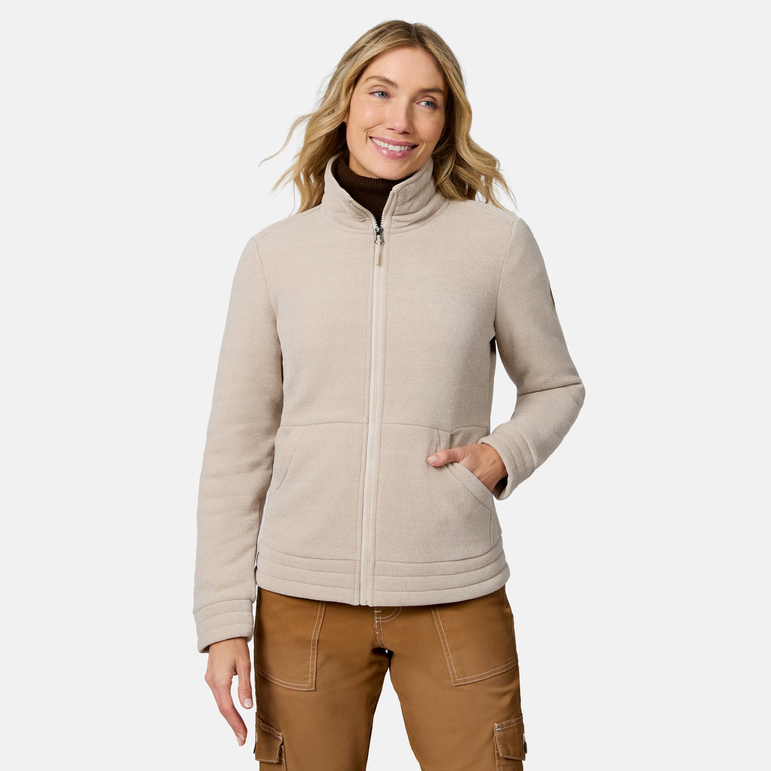 A woman wearing a light beige fleece zip-up jacket over a dark brown mock neck, paired with brown cargo pants, against a solid light gray background.