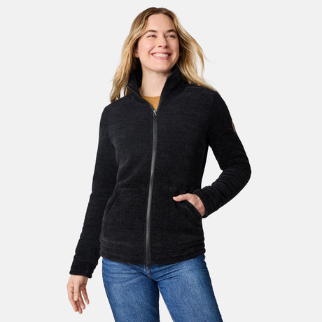 A woman wearing a dark charcoal zip-up fleece jacket and blue jeans, standing against a light gray studio background.