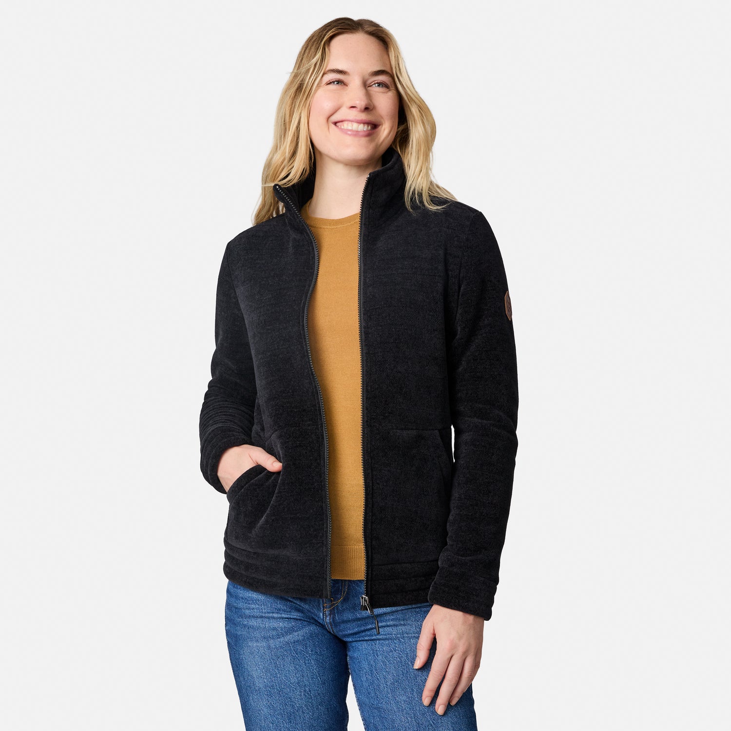 A woman modeling a dark charcoal zip-up fleece jacket over a mustard yellow top and blue jeans, photographed against a clean light gray studio background.