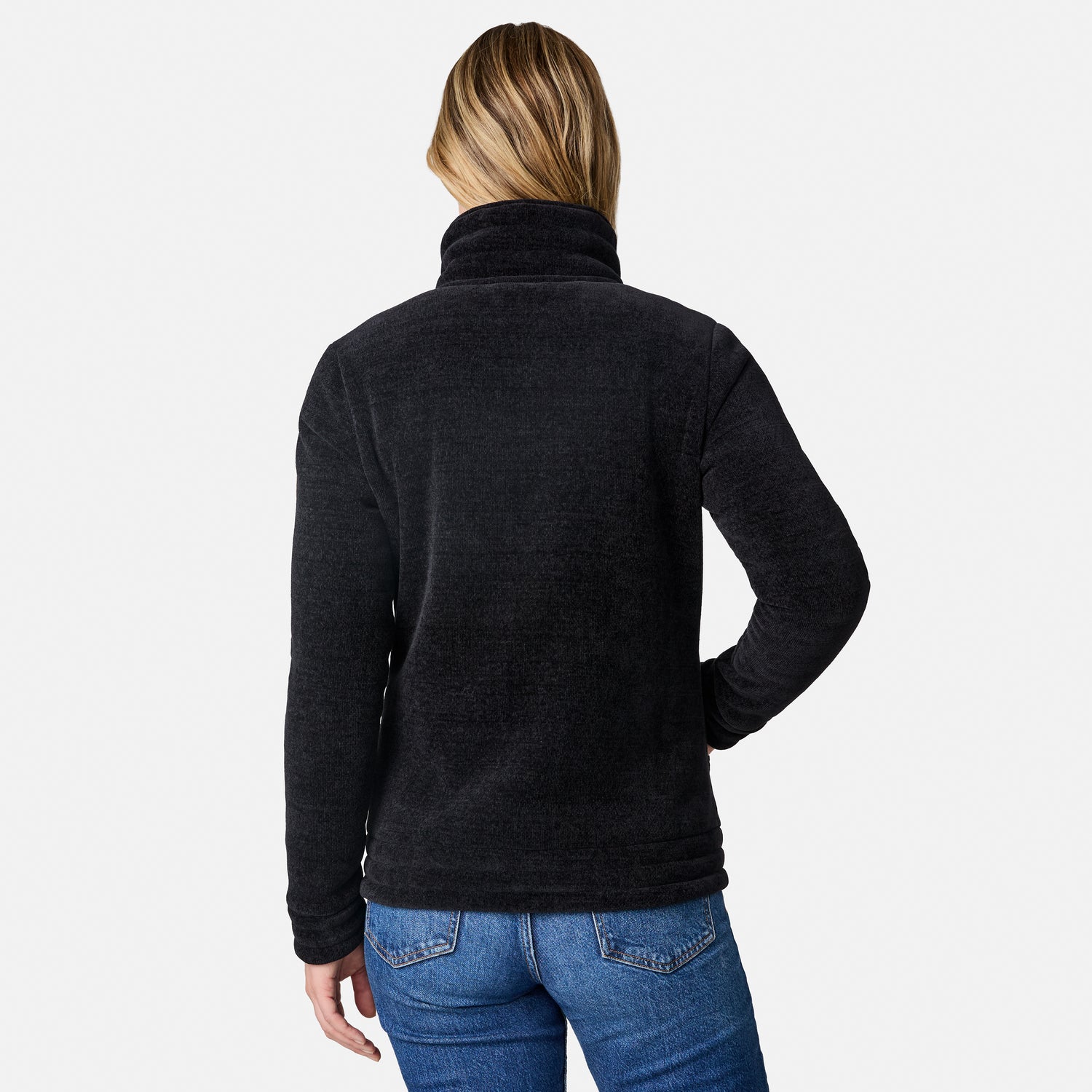 A woman viewed from the back wearing a dark charcoal heathered fleece pullover sweater and blue jeans against a light gray studio background.