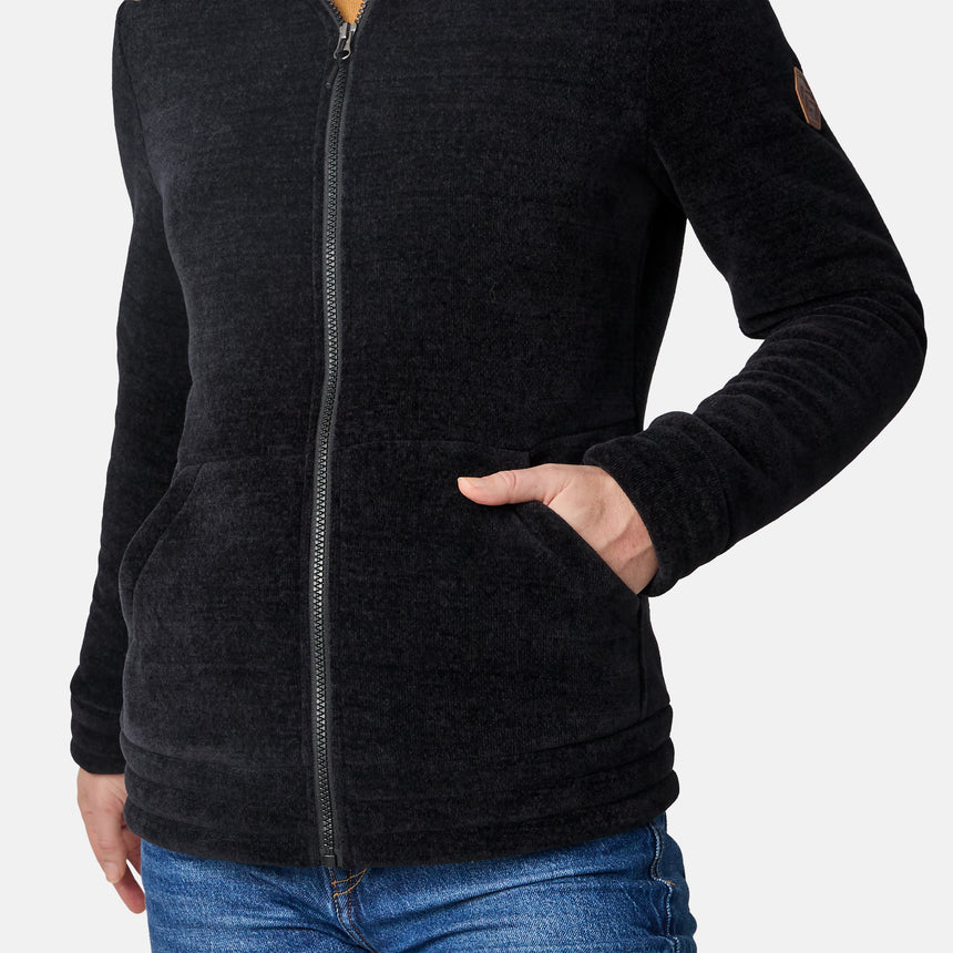 A close-up of a person wearing a black zip-up fleece jacket with a small brown patch on the sleeve, paired with blue jeans, against a light gray background.