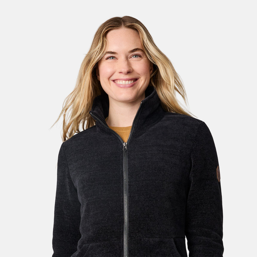 A smiling blonde woman wearing a dark charcoal zip-up fleece jacket over a mustard yellow shirt, photographed against a clean light gray background.