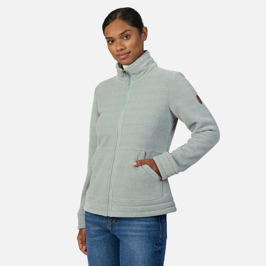 [Model is wearing size S.] A woman modeling a light green heather zip-up fleece jacket with a mock neck collar, paired with blue jeans, against a plain light gray studio background.