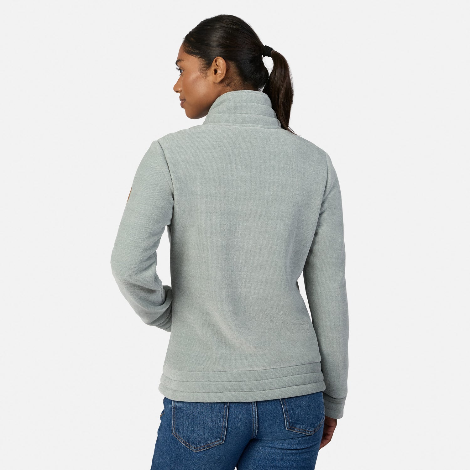 [Model is wearing size S.] A woman modeling the back of a light green heather zip-up fleece jacket with a mock neck collar, paired with blue jeans, against a plain light gray studio background.