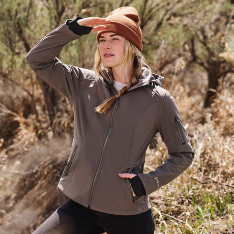 A woman outdoors in the sun shades her eyes while wearing a brown beanie and the Free Country Women's Aeris II Super Softshell® Jacket, a gray water resistant layer, with trees and dry grass behind her.