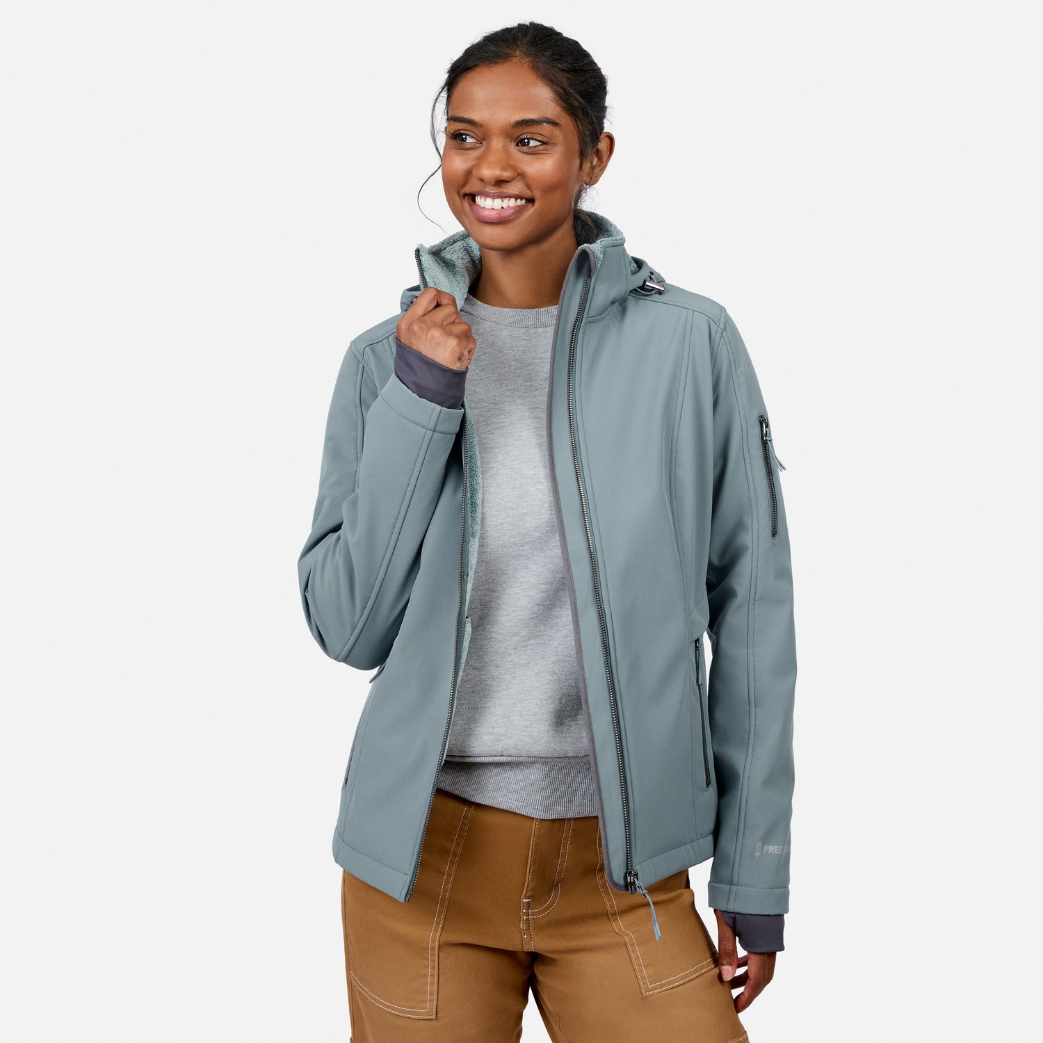 [Model is wearing size S.] A woman wearing a sage green hooded softshell jacket over a gray sweater and brown pants, standing against a light gray studio background.