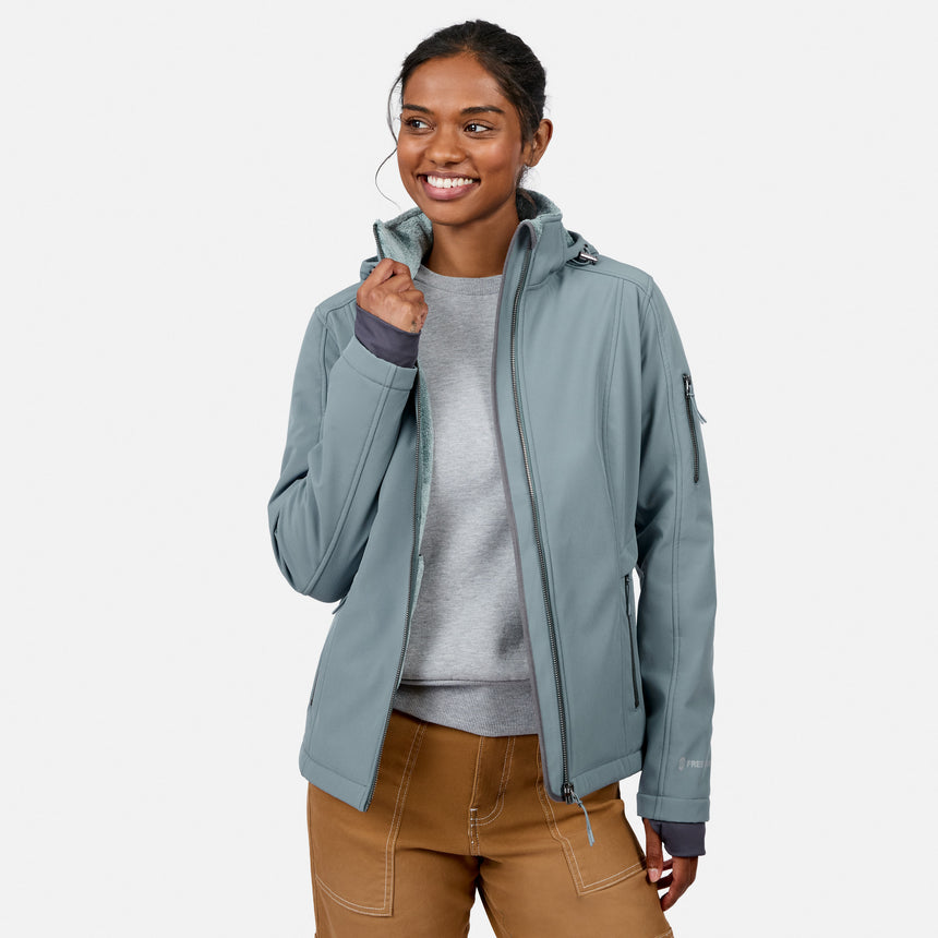 [Model is wearing size S.] A woman wearing a sage green hooded softshell jacket over a gray sweater and brown pants, standing against a light gray studio background.