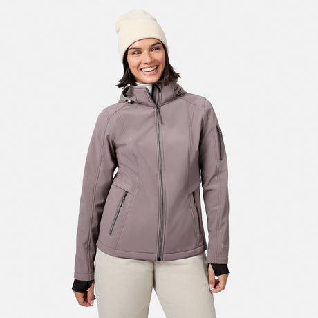 [Model is wearing size S.] A woman wearing a light taupe-gray hooded softshell jacket, a cream knit beanie, and light beige pants, smiling against a solid off-white background.