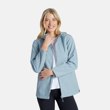 A smiling woman with blonde hair wears a light blue Women's Anorak Softshell Jacket by Clearance over a white shirt and black pants, standing against a plain light background.
