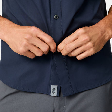 Close-up of a person buttoning the Free Country Men's Sedona Rip-Stop Short Sleeve Explorer Shirt, shown in dark navy with UPF protection and a small gray logo tag above gray pants. Only the hands and torso are visible.