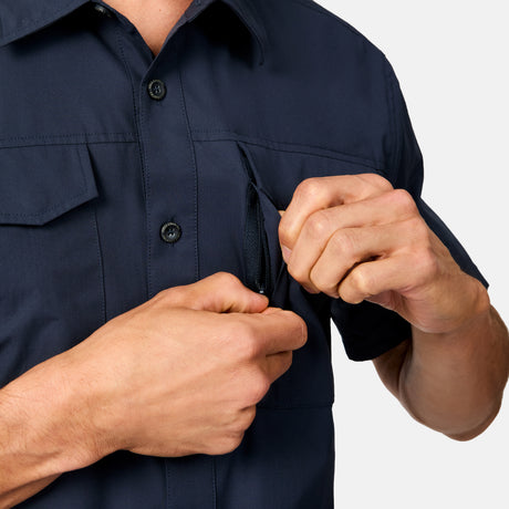 A person wearing the Free Country Men's Sedona Rip-Stop Short Sleeve Explorer Shirt in navy unzips a chest pocket with their right hand. Only the torso and hands are visible against a plain, light background.