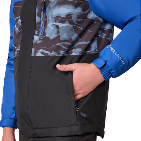 A person wears the Free Country Boys' Out of Bound Boarder Jacket with blue sleeves, a camouflage-patterned upper section, and black lower section. Their right hand is in a zippered pocket. The water-resistant sleeve reads "FREE COUNTRY.