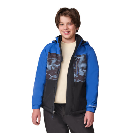 A smiling boy wears the Free Country Boys' Out of Bound Boarder Jacket, featuring a blue and black design with a camo chest, over a cream shirt and dark pants, standing against a white background.