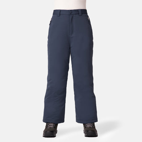 A person wears Free Country Kids' Padded Ski Pant in dark blue, featuring insulation, water resistance, zippered side pockets, and built-in snow gaiters. They're paired with black winter boots and a white ribbed shirt against a light gray background.