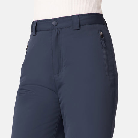 A child wears Free Country Kids' Padded Ski Pants in navy blue, featuring water resistance, zippered pockets, and belt loops, paired with a tucked-in light long-sleeve top. The image highlights the waist and upper thighs.