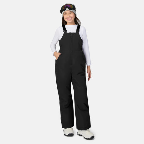 Smiling child in a black ski bib with straps, white long sleeve shirt gray beanie and goggles on the head