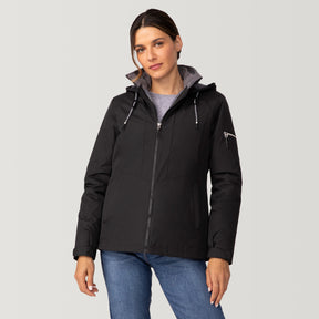 A woman wearing the Women's Back of Bell 3-in-1 Systems Jacket by Clearance in black stands against a plain light background. She styles the sustainable zip-up hooded jacket over a gray shirt and blue jeans, looking at the camera with a neutral expression.