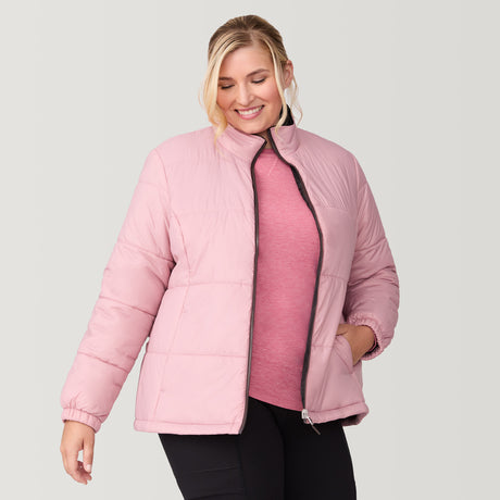 A woman with blonde hair smiles while wearing the Free Country Women's Plus Size Back of Bell 3-in-1 Systems Jacket in light pink over a pink top and black pants, standing against a plain light gray background.