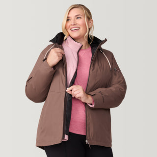 A smiling blonde woman models the Free Country Women's Plus Size Back of Bell 3-in-1 Systems Jacket in brown over a light pink zip-up and shirt, standing against a light gray background, holding the jacket open with one hand.