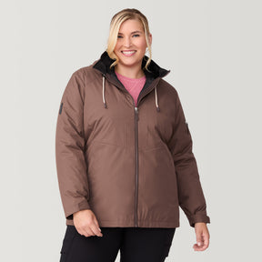 A smiling woman stands against a light background, wearing the Free Country Women's Plus Size Back of Bell 3-in-1 Systems Jacket in brown with a high collar and black drawstrings, over a pink top and black pants.