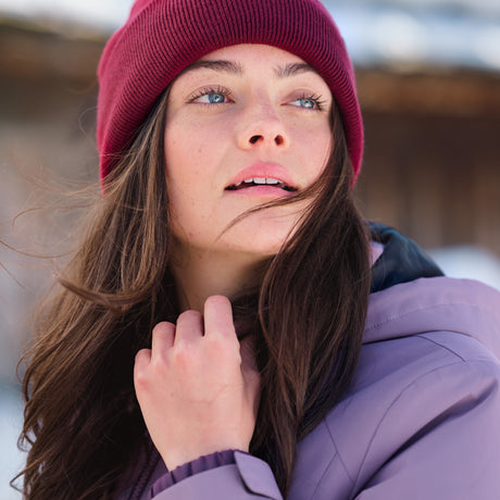 A woman in a maroon beanie and the Free Country Women's Back of Bell 3-in-1 Systems Jacket stands with her long brown hair blowing in the wind, gazing upward against a snowy, blurred winter backdrop.