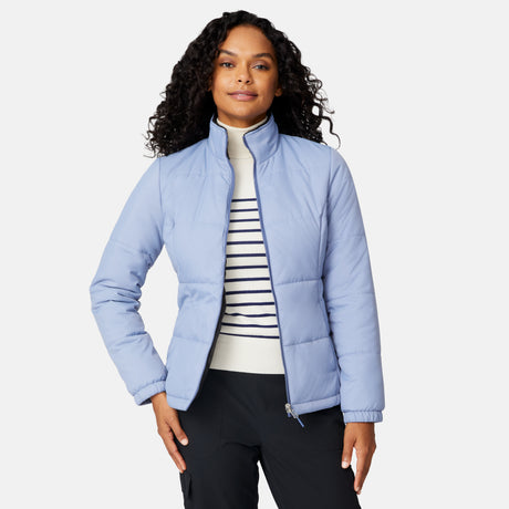 A woman with curly hair models the Free Country Women's Back of Bell 3-in-1 Systems Jacket in light blue over a white, navy-striped turtleneck and black pants, standing against a plain white background.