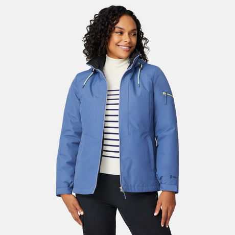 A woman with curly hair smiles while wearing the Free Country Women's Back of Bell 3-in-1 Systems Jacket in blue over a striped white turtleneck, standing against a plain, light background with her hands at her sides.