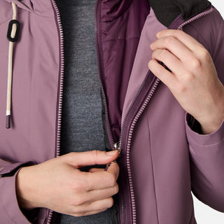 A person is shown zipping up a mauve Women's Back of Bell 3-in-1 Systems Jacket by Free Country over a gray shirt. The photo highlights the water-resistant, sustainable jacket's hands and torso, with the jacket partially unzipped.