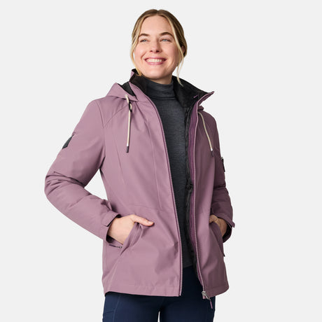 A woman smiles while wearing the Free Country Women's Back of Bell 3-in-1 Systems Jacket in light purple with black inner lining, paired with a dark turtleneck and pants. She stands against a plain light background, hands in her jacket pockets.