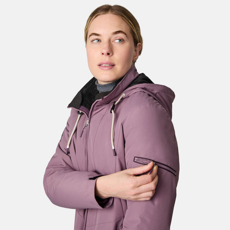 A light-haired woman, hair tied back, in a mauve Free Country Women's Back of Bell 3-in-1 Systems Jacket unzips a small upper left sleeve pocket, looking slightly to the side against a plain white background.