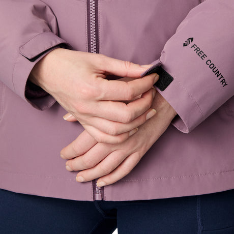 Close-up of a person adjusting the Velcro cuff on a mauve Free Country Women's Back of Bell 3-in-1 Systems Jacket, with their other hand resting on top; the brand name and logo are visible on the sleeve.