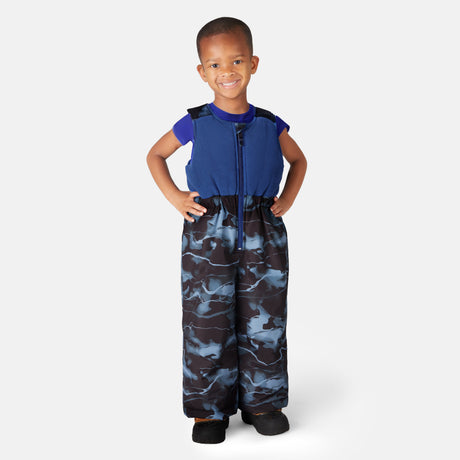 A smiling child wears the Free Country Toddler Boys' Out of Bound Jacket and Bib Pant Ski Set, featuring a blue short-sleeve shirt and black, blue, and gray camouflage, standing with hands on hips against a plain light gray background.
