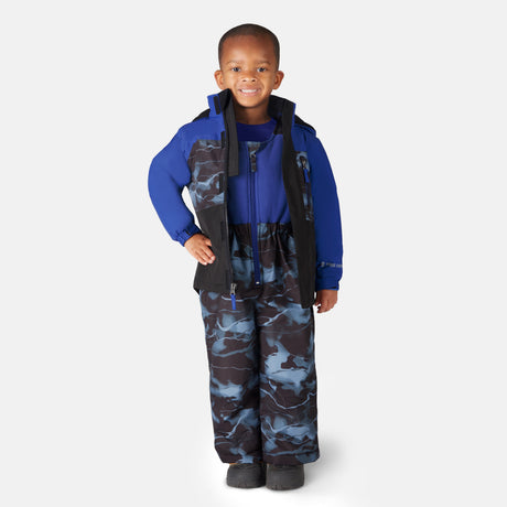 A young child smiles while standing against a plain light background, wearing the Free Country Toddler Boys' Out of Bound Jacket and Bib Pant Ski Set with matching blue and black camo print and black boots.