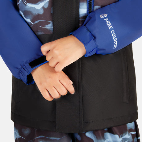 A child adjusts the Velcro strap on a blue and black Toddler Boys' Out of Bound Jacket by Free Country, paired with matching camouflage bib pants—part of a water-resistant ski set.