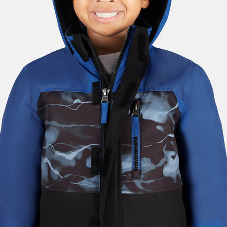 A child smiles wearing the Free Country Toddler Boys' Out of Bound Jacket and Bib Pant Ski Set. The blue, black, and gray water-resistant set features a zipper and Velcro closures, perfect for winter fun. Background is plain white.