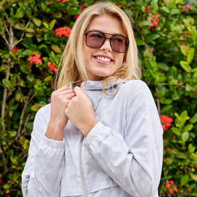 A smiling woman in sunglasses wears the Clearance Women's Outland Windshear Jacket, light gray and wind resistant, standing outdoors among green foliage and red flowers.