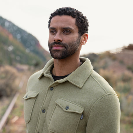 Man wearing a tan button up coat standing outdoors with mountains in the background
