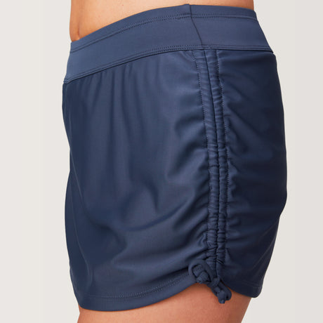 A close-up side view shows a woman wearing the Free Country Women's Side Shirred Swim Skirt, featuring navy blue color, adjustable side ties, a built-in brief, and ruched detail against a light background that highlights its stylish design.